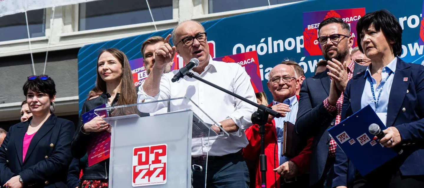 Polityk przemawia z mikrofonem na podium podczas wiecu, otoczony innymi uczestnikami i plakatami w tle.