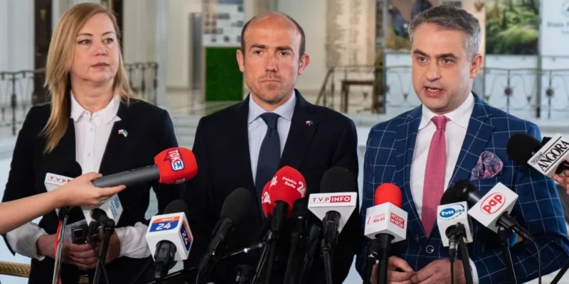 Trzech polityków stoi podczas konferencji prasowej, otoczonych licznymi mikrofonami reporterów w wnętrzu budynku.