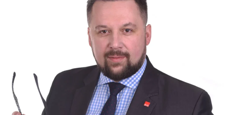 Portret mężczyzny w granatowym garniturze, niebieskiej koszuli w kratkę i granatowym krawacie, trzymającego okulary, z czerwonym PINem SLD na klapie i kwiecistą poszetką w kieszeni na białym tle.