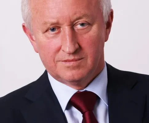 Portret starszego mężczyzny w granatowym garniturze i bordowym krawacie na jasnym tle.