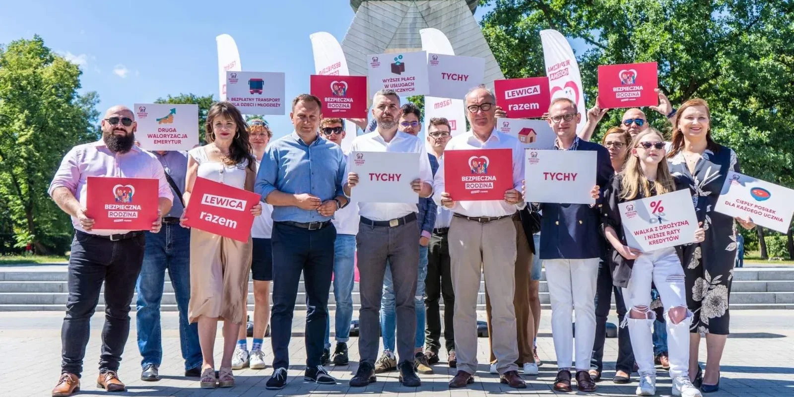 Grupa osób stoi na placu, trzymając plakaty z hasłami „Bezpieczna Rodzina” i „Lewica razem” podczas plenerowej akcji promującej trasę Bezpieczna Rodzina, w tle widoczny jest pomnik.