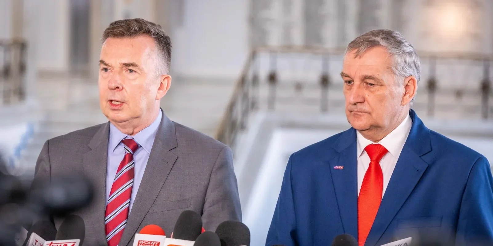 Dwóch mężczyzn w garniturach przemawia do mediów podczas konferencji prasowej, stojąc przy licznych mikrofonach.