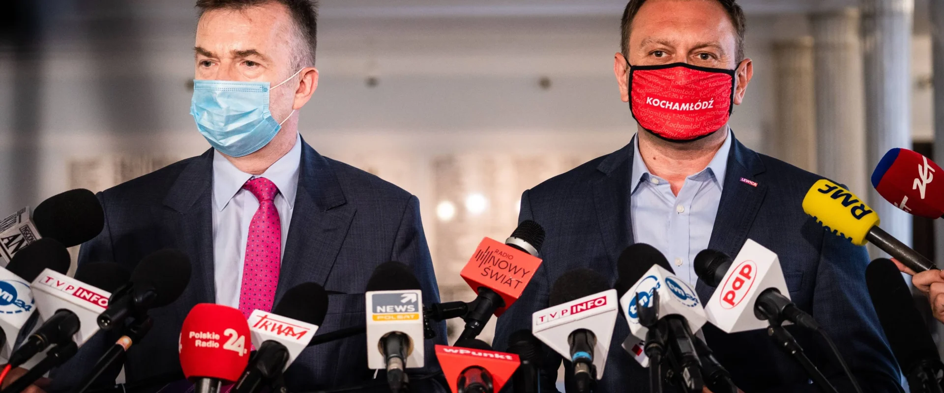 Dwóch polityków Lewicy podczas konferencji prasowej otoczonych licznymi mikrofonami, jeden w niebieskiej maseczce, drugi w czerwonej z napisem „Kocham Łódź”.
