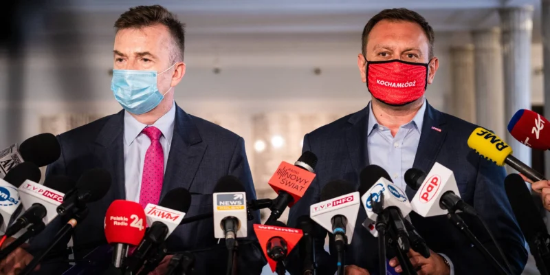 Dwóch polityków Lewicy podczas konferencji prasowej otoczonych licznymi mikrofonami, jeden w niebieskiej maseczce, drugi w czerwonej z napisem „Kocham Łódź”.