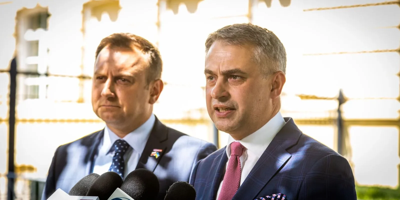 Dwóch polityków w garniturach przemawia do dziennikarzy podczas konferencji prasowej, stojąc przed mikrofonami na tle budynku.