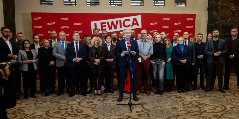 Grupa polityków Nowej Lewicy podczas konferencji prasowej w sprawie umowy koalicyjnej i realizacji programu wyborczego, stojąca przed czerwonym banerem LEWICA, z centralnym mówcą przy mikrofonie.