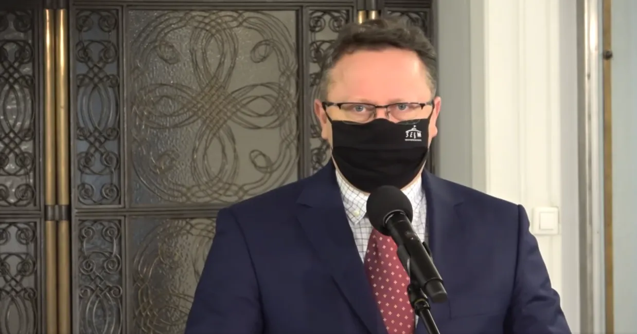 Mężczyzna w garniturze i maseczce stoi przy mikrofonie podczas konferencji prasowej.