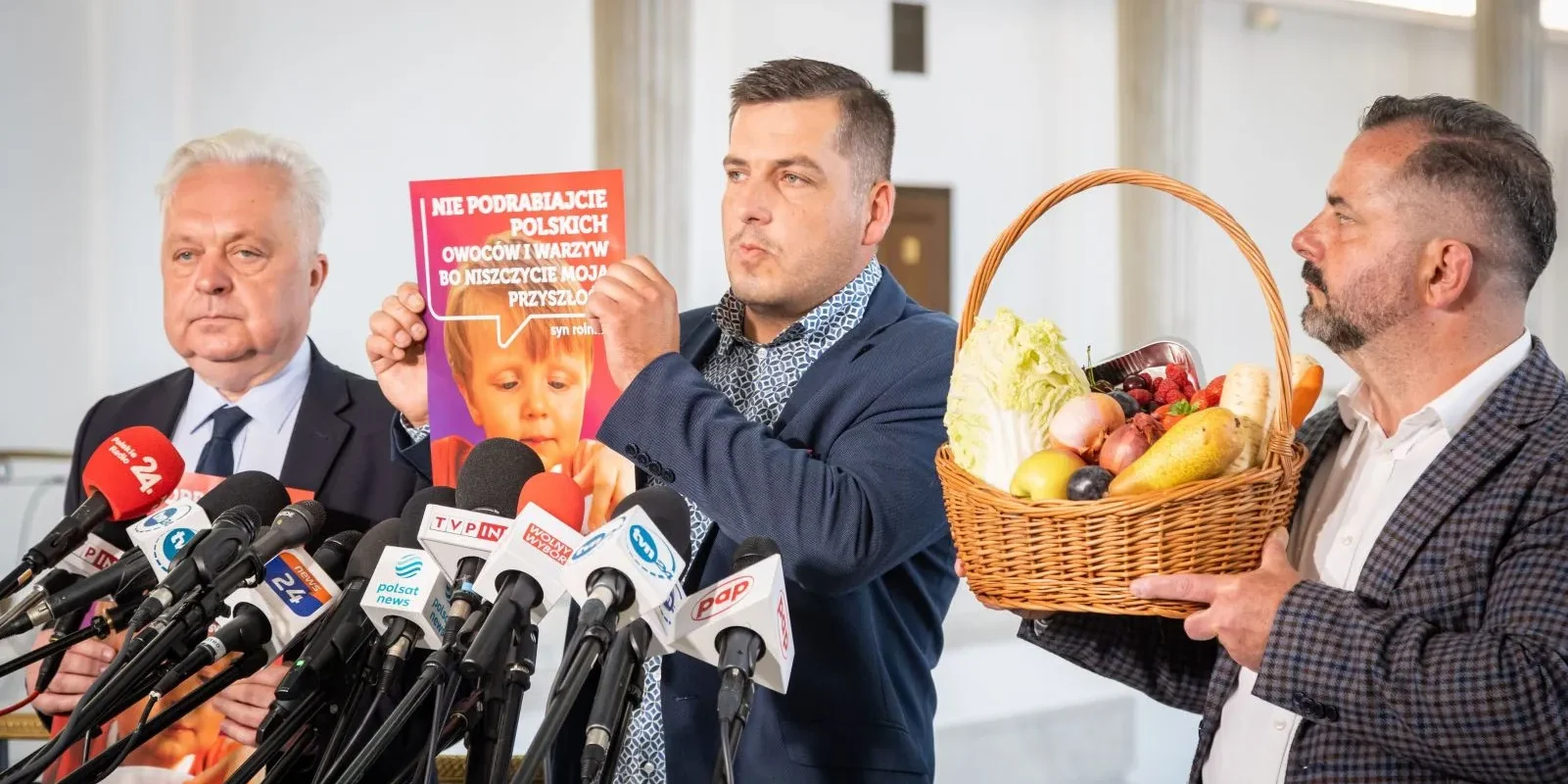 Na zdjęciu przedstawieni są mężczyźni podczas konferencji prasowej, z jednym z nich trzymającym plakat z hasłem o ochronie polskich owoców i warzyw, a obok widoczny jest kosz pełen świeżych produktów.