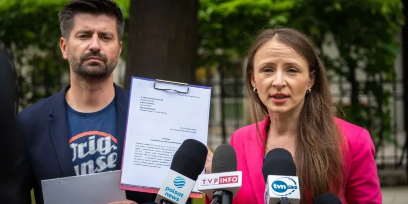 Dwóch dorosłych podczas konferencji prasowej na zewnątrz; kobieta w różowej marynarce przemawia do mikrofonów, mężczyzna trzyma teczkę z dokumentem, a w tle są drzewa.