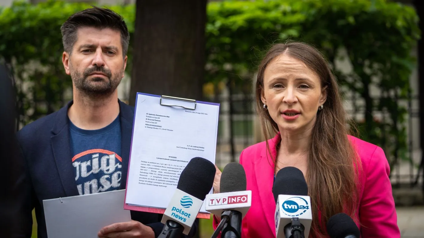 Dwóch dorosłych podczas konferencji prasowej na zewnątrz; kobieta w różowej marynarce przemawia do mikrofonów, mężczyzna trzyma teczkę z dokumentem, a w tle są drzewa.