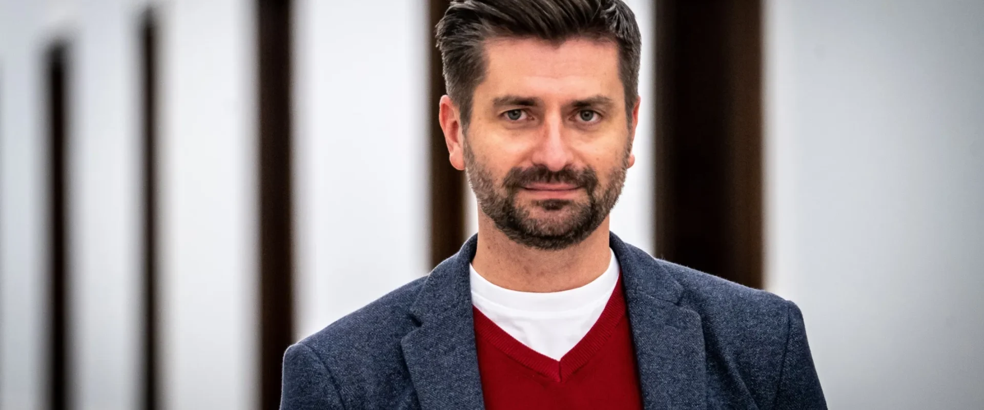Mężczyzna w granatowej marynarce i czerwonym swetrze stoi na tle jasnej ściany z ciemnymi drzwiami.