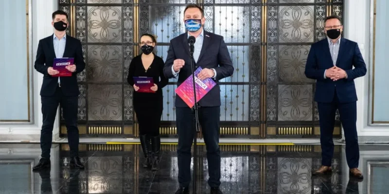 Czterech osób w garniturach i maseczkach stoi na błyszczącym marmurowym foyer przed ozdobnymi drzwiami; mężczyzna w środku przemawia do mikrofonu, trzymając kolorowe ulotki.