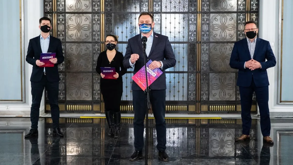 Czterech osób w garniturach i maseczkach stoi na błyszczącym marmurowym foyer przed ozdobnymi drzwiami; mężczyzna w środku przemawia do mikrofonu, trzymając kolorowe ulotki.