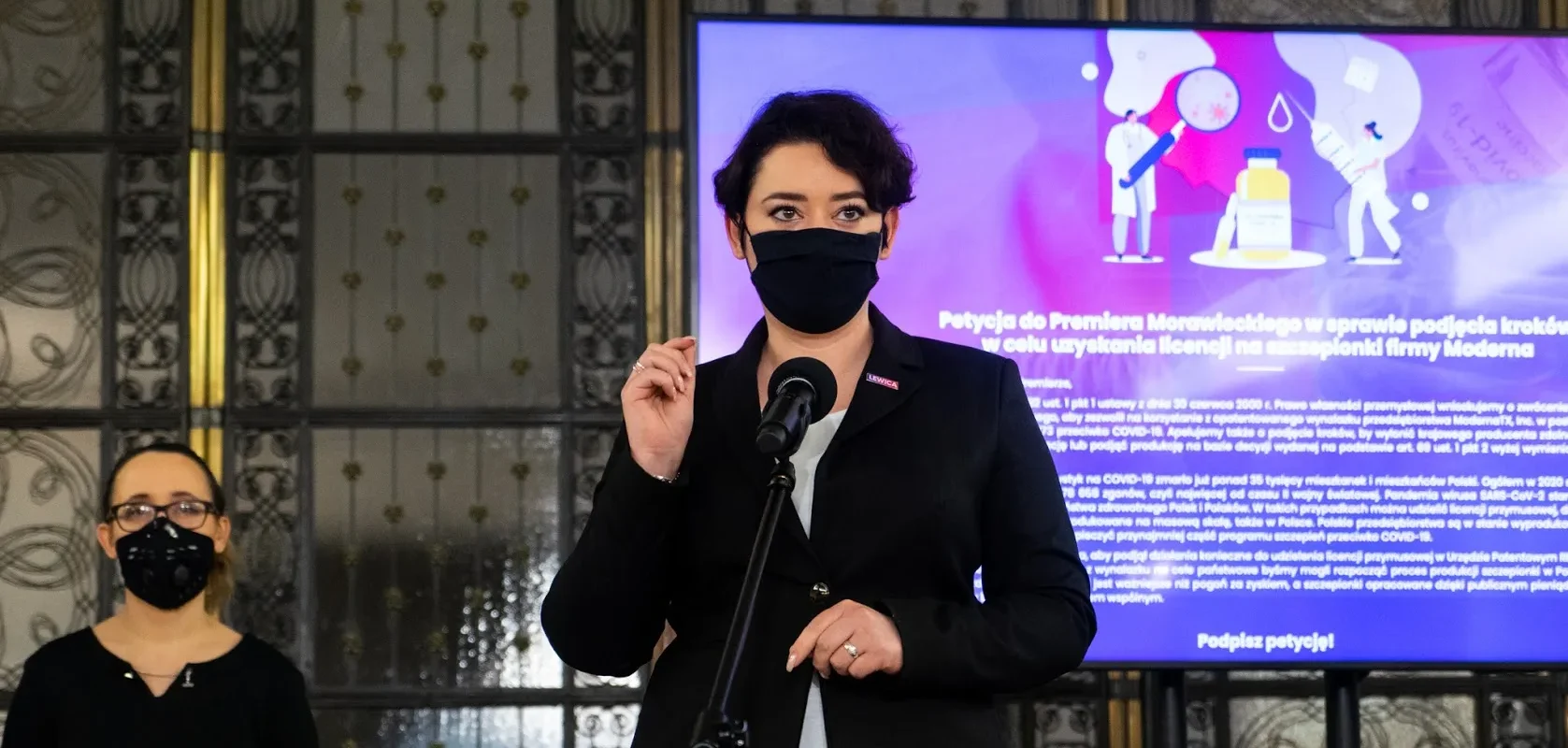 Kobieta w czarnym żakieciku i czarnej maseczce stoi przy mikrofonie podczas konferencji; w tle duży ekran z ilustracją medyczną w fioletowych barwach, a z lewej strony stoi druga osoba w maseczce.