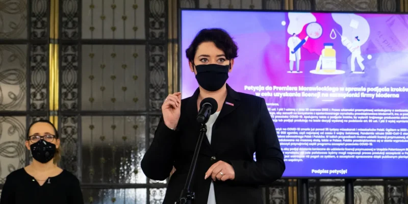 Kobieta w czarnym żakieciku i czarnej maseczce stoi przy mikrofonie podczas konferencji; w tle duży ekran z ilustracją medyczną w fioletowych barwach, a z lewej strony stoi druga osoba w maseczce.