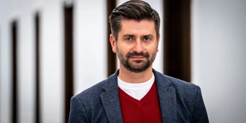 Na zdjęciu widoczny jest mężczyzna w eleganckim ubraniu, stojący na tle jasnej ściany z uchylonymi drzwiami.