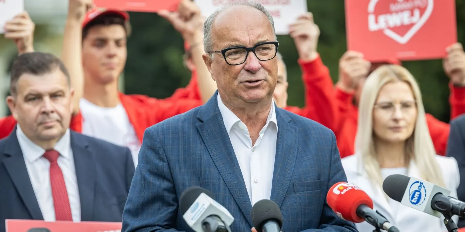 Mężczyzna w granatowym garniturze i czarnych okularach przemawia do mikrofonów podczas konferencji prasowej; w tle grupa osób w czerwonych koszulkach trzyma plakaty.