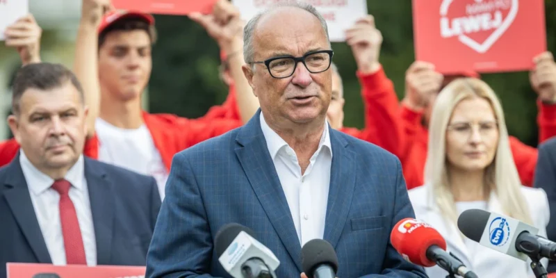 Mężczyzna w granatowym garniturze i czarnych okularach przemawia do mikrofonów podczas konferencji prasowej; w tle grupa osób w czerwonych koszulkach trzyma plakaty.