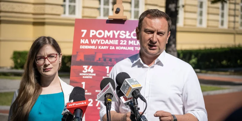 Mężczyzna i kobieta stoją przed mikrofonami podczas konferencji prasowej, obok czerwonego plakatu z informacją o 22 mln zł dziennej kary za Kopalnię Turów.