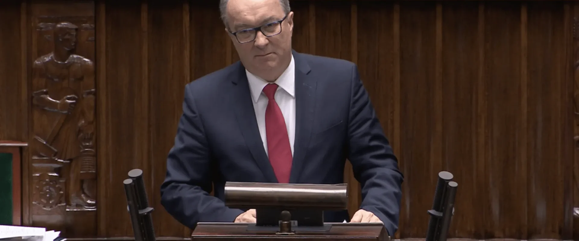 Na zdjęciu przedstawiono mężczyznę w garniturze, stojącego za mównicą w sali obrad, z poważnym wyrazem twarzy.