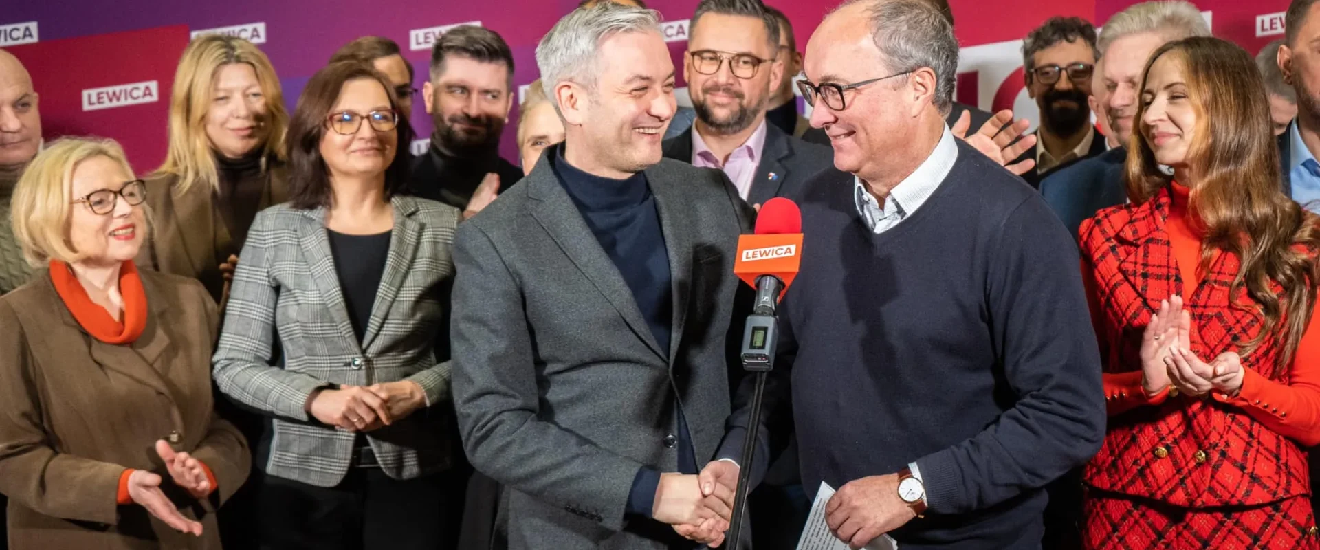 Grupa osób na tle różowo-fioletowego tła z logo Lewica, na pierwszym planie dwóch mężczyzn ściska sobie dłonie przy mikrofonie z napisem LEWICA, reszta uczestników uśmiecha się i klaszcze.