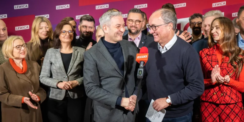 Grupa osób na tle różowo-fioletowego tła z logo Lewica, na pierwszym planie dwóch mężczyzn ściska sobie dłonie przy mikrofonie z napisem LEWICA, reszta uczestników uśmiecha się i klaszcze.