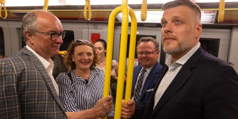 Grupa pasażerów w eleganckich ubraniach stoi w pociągu, trzymając się żółtych uchwytów i rozmawiając.