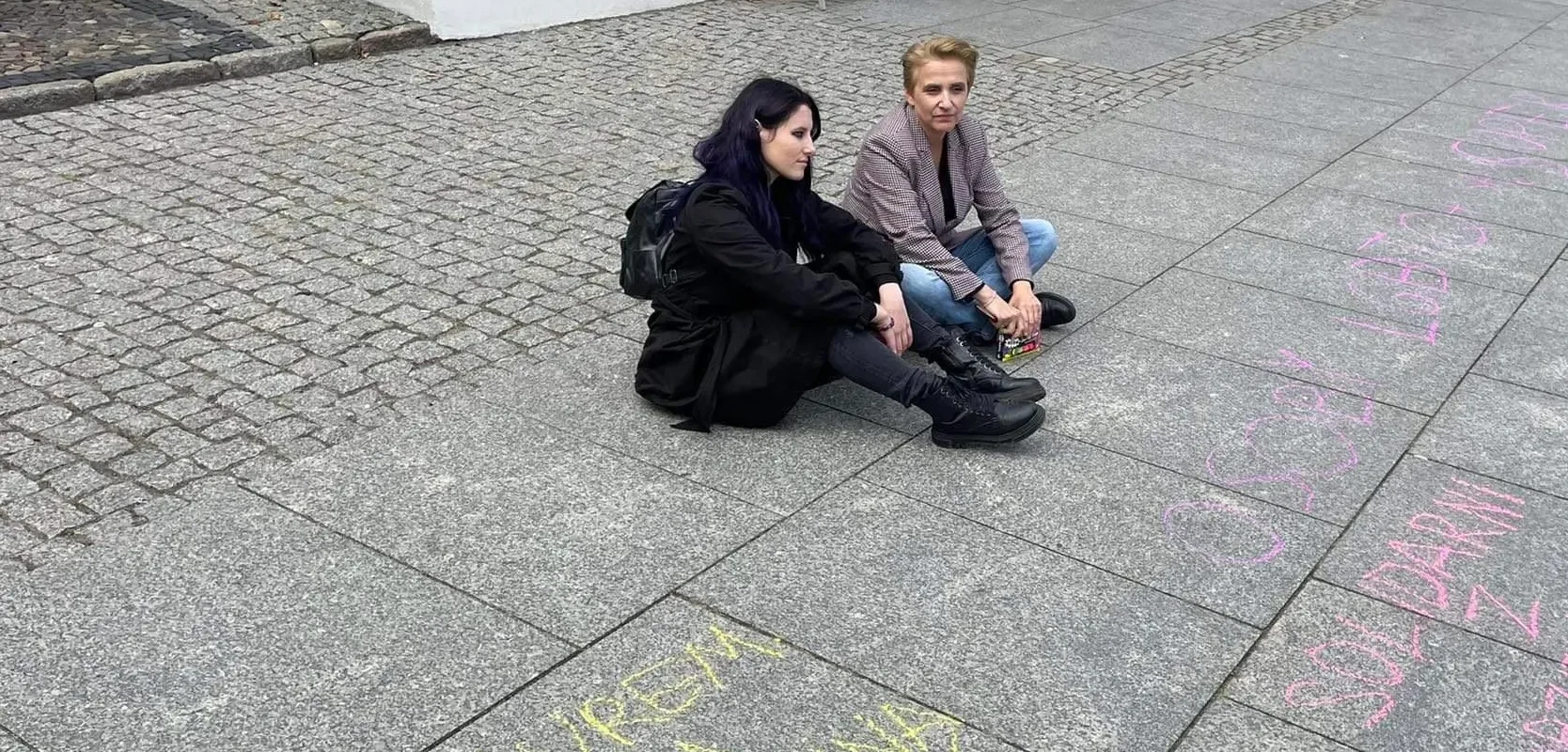 Na zdjęciu widać dwie osoby siedzące na chodniku, obok nich są kolorowe napisy na nawierzchni.
