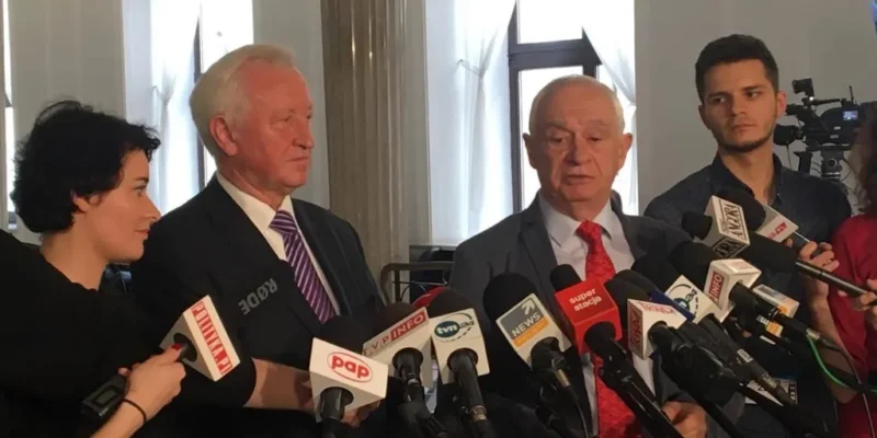 Polityk i współpracownicy stoją przed licznymi mikrofonami podczas konferencji prasowej o tym, że rząd nie sięga po środki UE na pomoc poszkodowanym w nawałnicach.