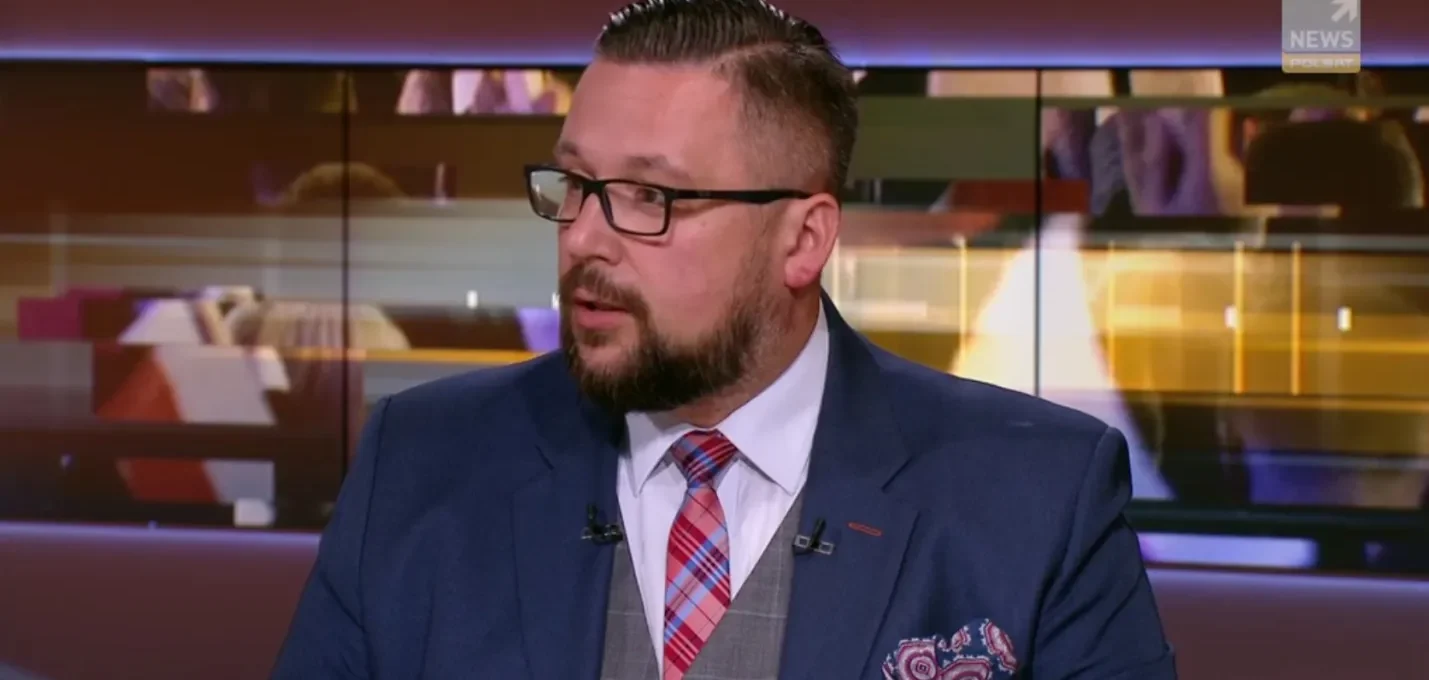 Mężczyzna w granatowym garniturze z okularami, białą koszulą i czerwonym w kratę krawatem siedzi w studiu telewizyjnym.