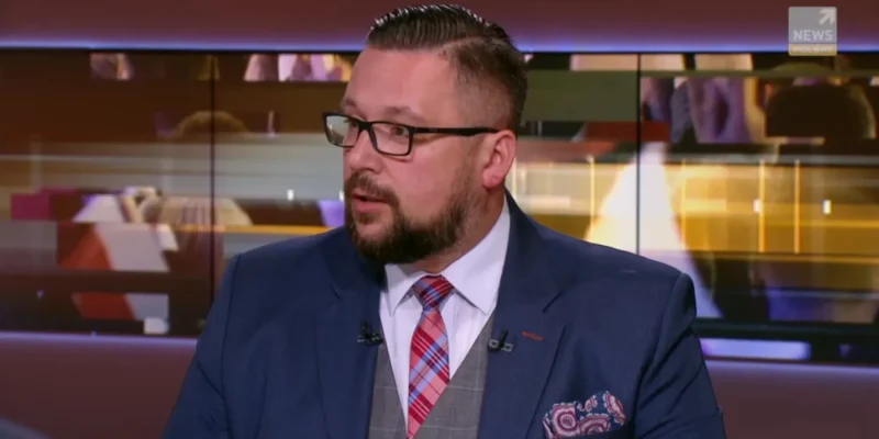 Mężczyzna w granatowym garniturze z okularami, białą koszulą i czerwonym w kratę krawatem siedzi w studiu telewizyjnym.