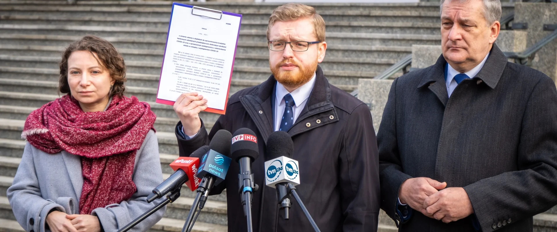 Na zdjęciu trzech polityków stoi przed schodami; jeden z nich trzyma dokument, podczas gdy pozostałe osoby uważnie go słuchają.