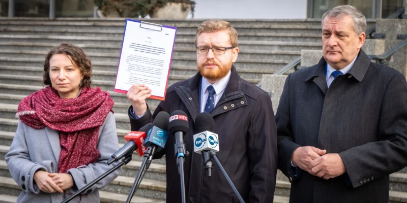 Na zdjęciu trzech polityków stoi przed schodami; jeden z nich trzyma dokument, podczas gdy pozostałe osoby uważnie go słuchają.