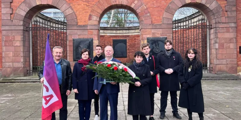 Grupa ludzi stoi przed historyczną bramą, trzymając wieńce i flagi, w celu upamiętnienia ważnej chwili.