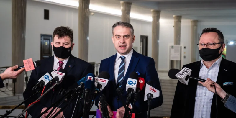 Trzech mężczyzn w garniturach i maseczkach stoi podczas konferencji prasowej otoczonych mikrofonami, omawiając wyciek danych z polskiego wojska.