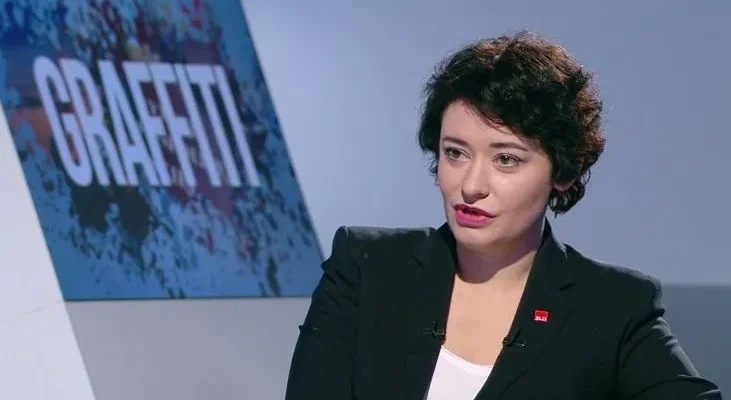 Kobieta w eleganckim garniturze wypowiadająca się w studiu telewizyjnym podczas wywiadu, z planszą "GRAFFITI" w tle.