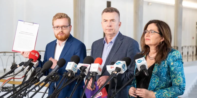 Trzy osoby w garniturach stoją na konferencji prasowej, otoczone licznymi mikrofonami.