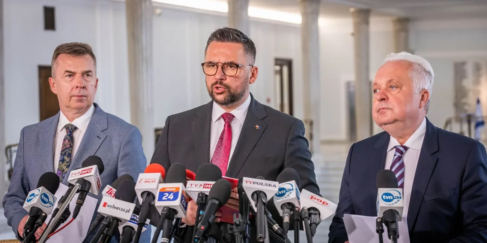 Na zdjęciu widać trzech mężczyzn przy mównicy z mikrofonami, którzy reprezentują lewicową partię i domagają się kontroli NIK rynku nawozów w Polsce.