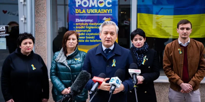 Grupa osób podczas konferencji prasowej na zewnątrz budynku, z banerem „Pomoc dla Ukrainy” i flagą Ukrainy w tle, wszyscy mają żółto-niebieskie wstążki.