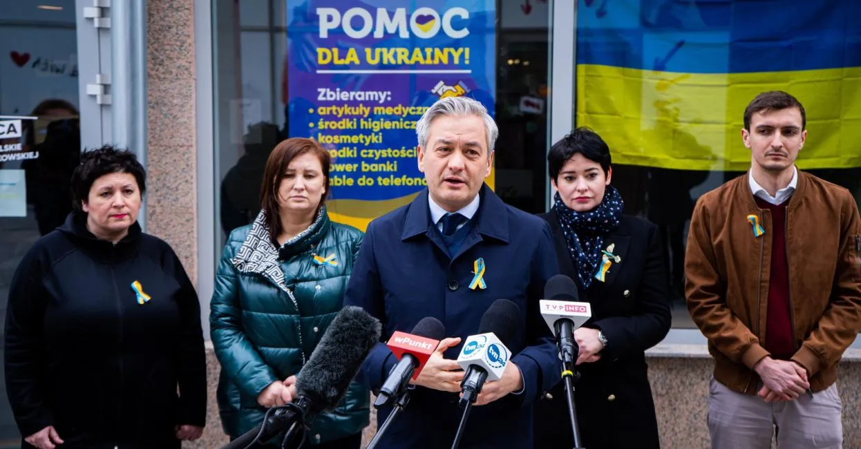 Grupa osób podczas konferencji prasowej na zewnątrz budynku, z banerem „Pomoc dla Ukrainy” i flagą Ukrainy w tle, wszyscy mają żółto-niebieskie wstążki.