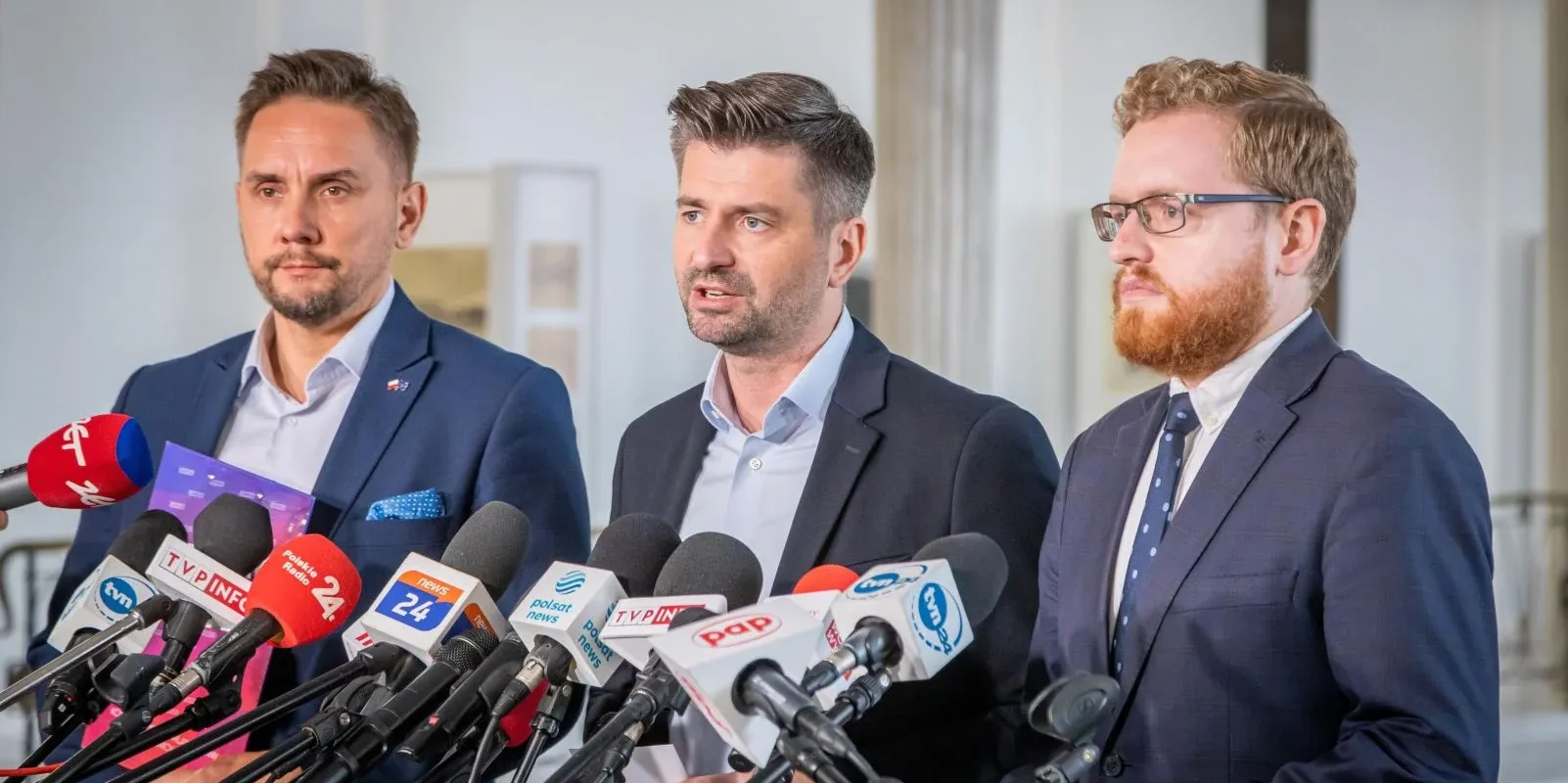 Trzech mężczyzn w garniturach stoi przed mikrofonami, prowadząc konferencję prasową w jasnym wnętrzu.