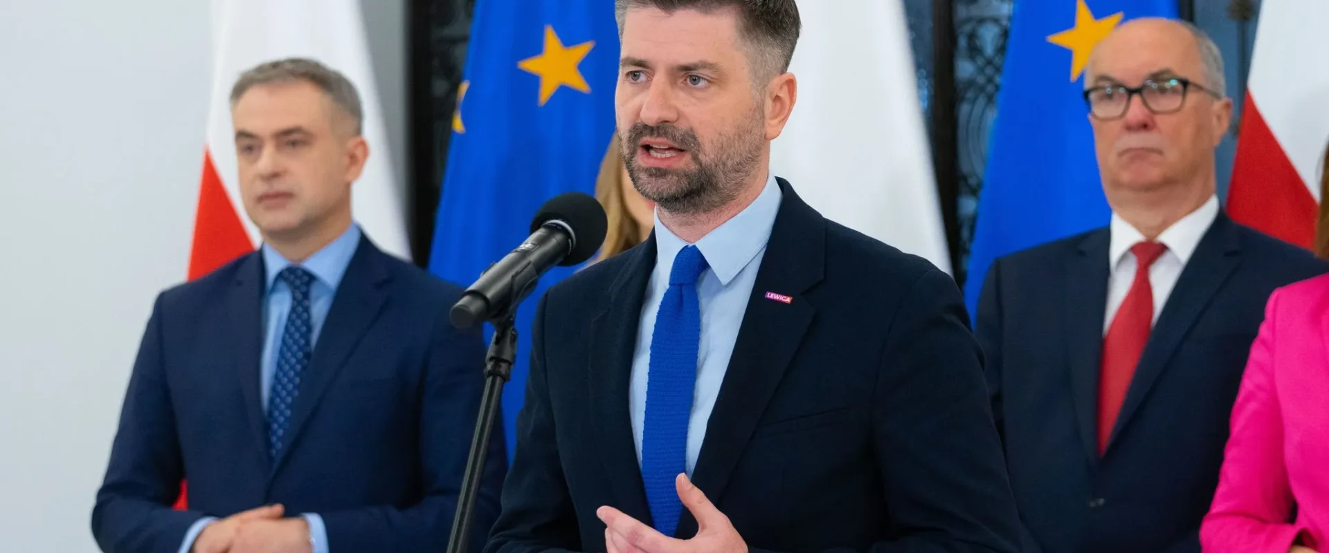 Polityk Lewicy w garniturze przemawia do mikrofonu podczas konferencji prasowej, z flagami UE w tle.