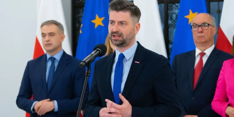 Polityk Lewicy w garniturze przemawia do mikrofonu podczas konferencji prasowej, z flagami UE w tle.