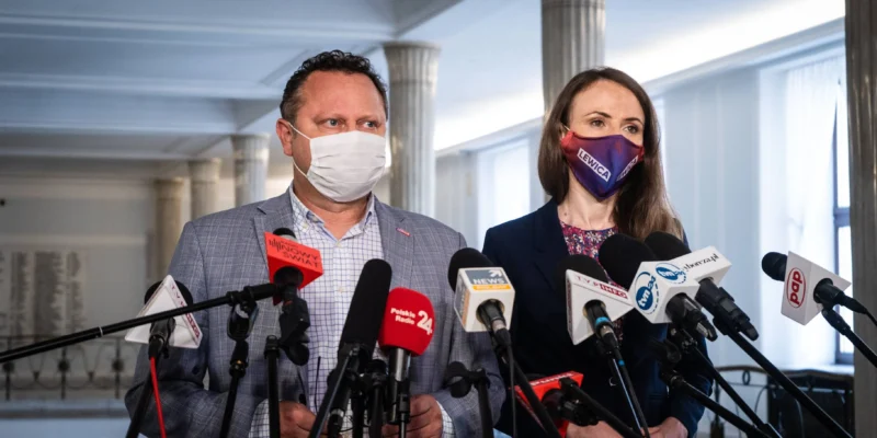 Dwóch dorosłych stoi przy konferencji prasowej z licznymi mikrofonami; mężczyzna w szarej marynarce i białej masce oraz kobieta w granatowej marynarce i maseczce z napisem Lewica, w tle kolumny i wnętrze budynku.