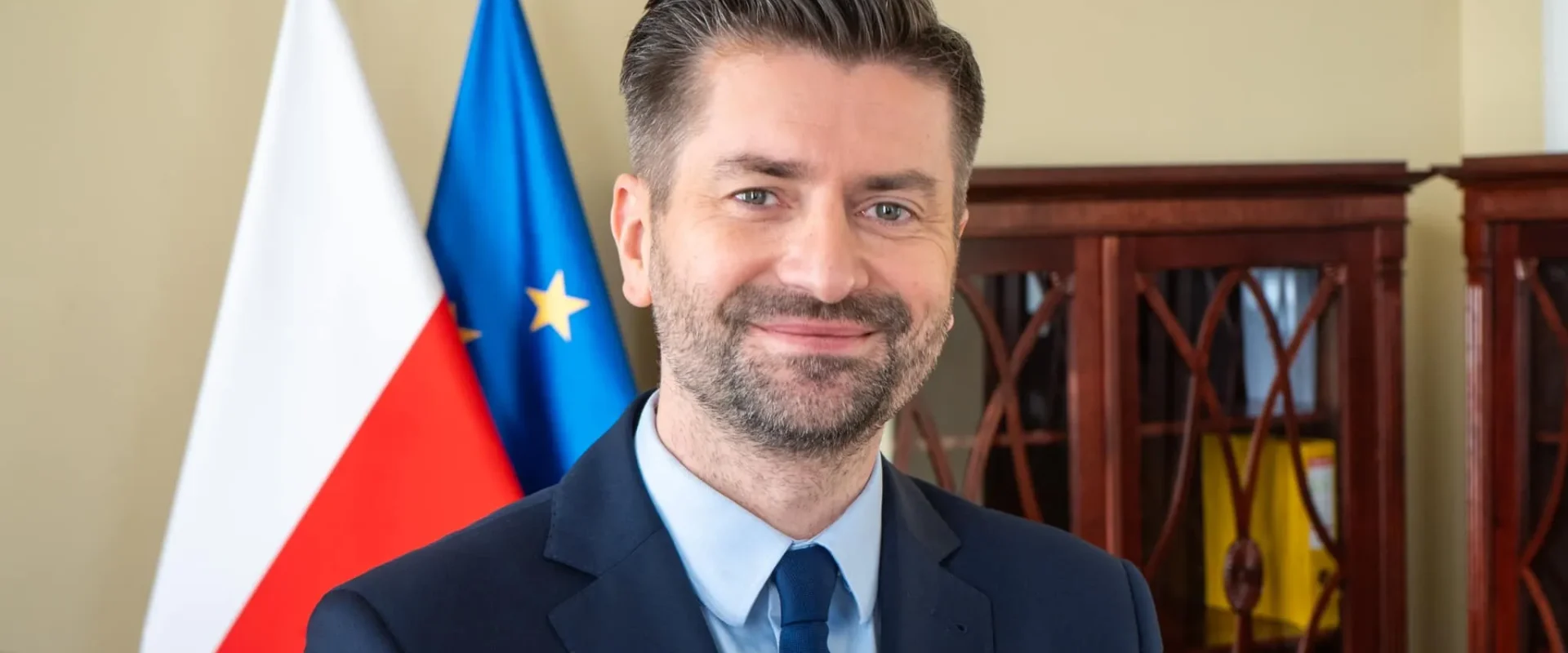 Mężczyzna w garniturze stoi na tle flag Polski i Unii Europejskiej, jako polski kandydat na komisarza UE.