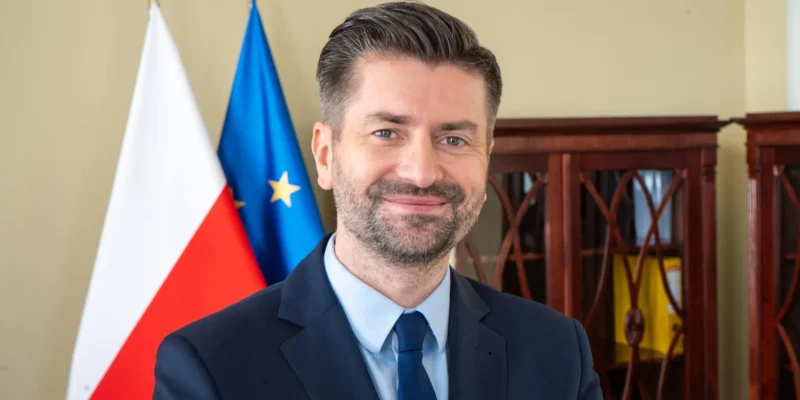 Mężczyzna w garniturze stoi na tle flag Polski i Unii Europejskiej, jako polski kandydat na komisarza UE.