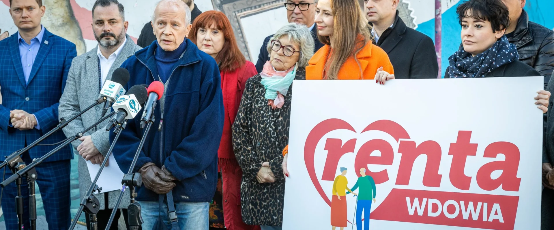 Grupa osób na konferencji prasowej trzyma plakat z napisem „Renta Wdowia” i stoi wśród mikrofonów, na tle kolorowego muralu.