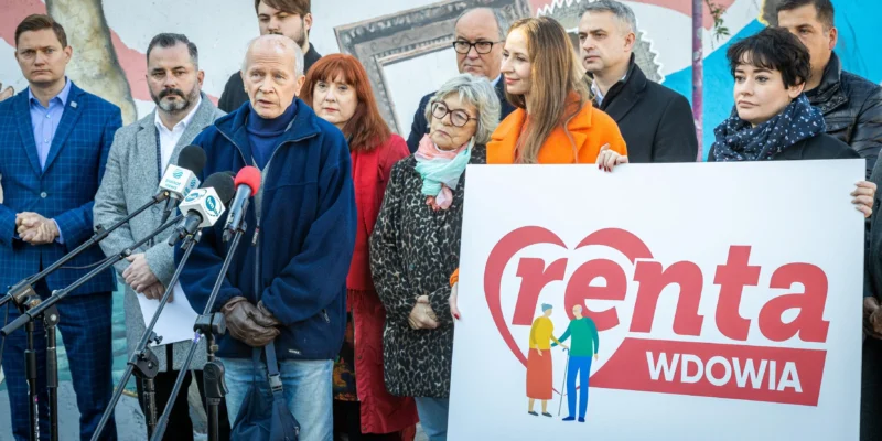 Grupa osób na konferencji prasowej trzyma plakat z napisem „Renta Wdowia” i stoi wśród mikrofonów, na tle kolorowego muralu.