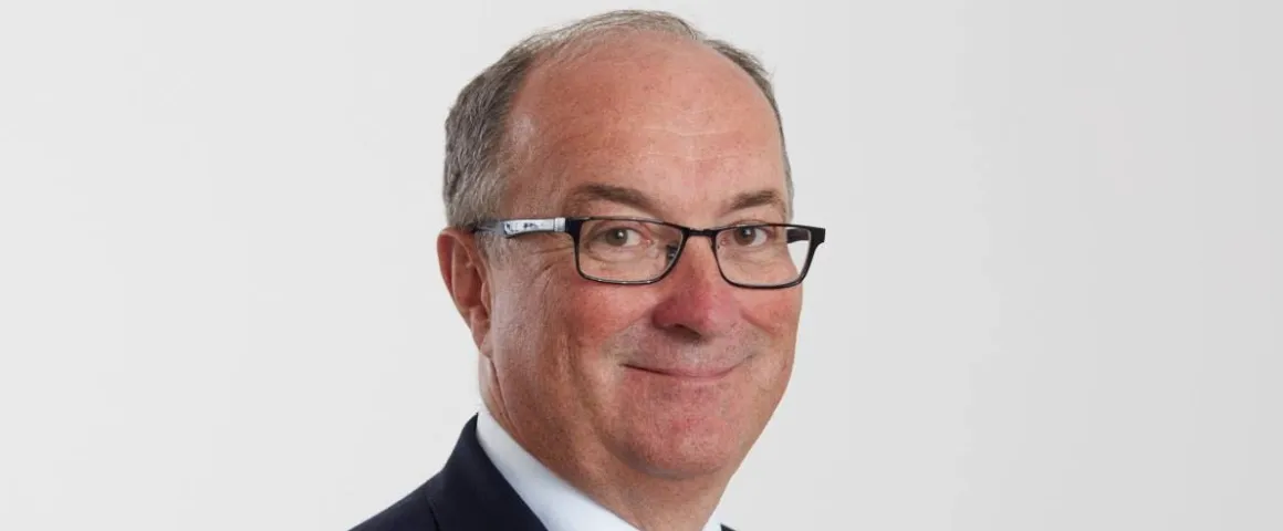 Portret mężczyzny w okularach, uśmiechniętego, w garniturze na jasnym tle.