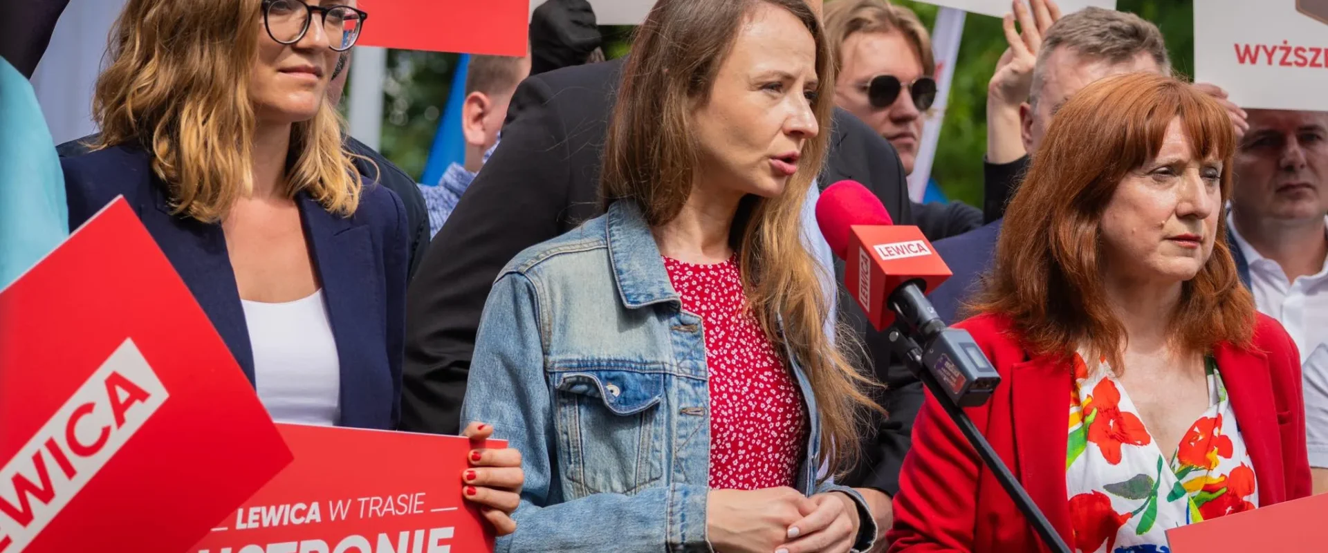 Grupa osób na demonstracji politycznej trzyma czerwone plakaty Lewica; na pierwszym planie kobieta w jeansowej kurtce wygłasza przemówienie do mikrofonu, a obok niej stoi kobieta w czerwonej marynarce.